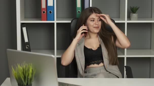 Joven mujer de negocios hermosa de apariencia caucásica con ojos marrones trabaja en su escritorio, sosteniendo un teléfono en sus manos hablando con los clientes — Vídeo de stock