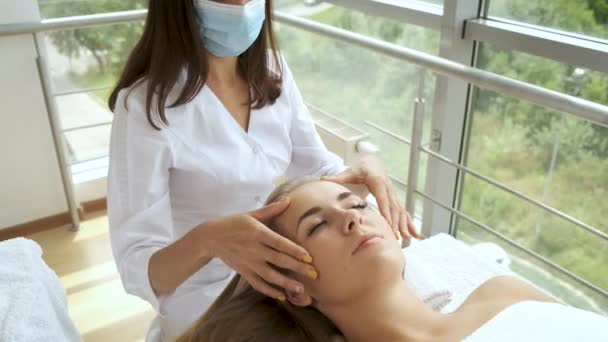 Een vrouw met een masker massages de patiënten hoofd in een moderne spa met panoramische ramen. Gezondheid en zelfzorg. Ontspanning — Stockvideo