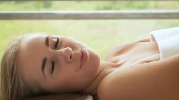 Een blonde blanke vrouw ligt in een spa met panoramische ramen en kaarsen en bloemen op de tafel. Ontspanning en gezondheid — Stockvideo