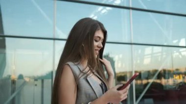 Beyaz takım elbiseli güzel bir kadın elinde telefonla dışarıda yürüyor ve kulaklıkla müzik dinliyor.