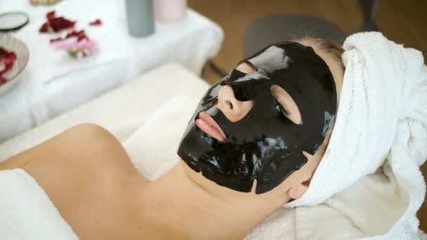 Женщина лежит в спа-салоне с черной маской на лице. Омоложение и здоровье кожи — стоковое видео