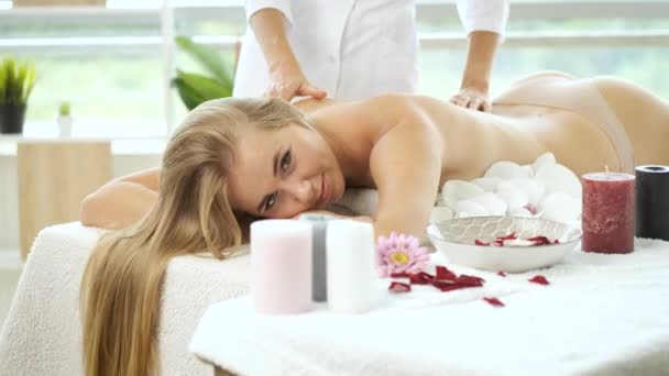 Masaje de espalda sobre mesa de masaje con flores y velas a hermosa chica rubia en el moderno centro de spa. Rehabilitación y salud. Relájate. — Vídeo de stock