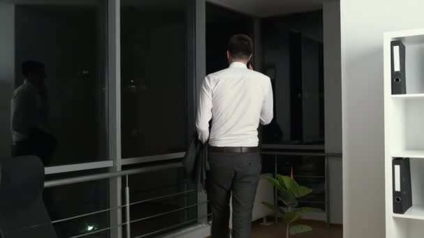 スタイリッシュなスーツの黒い髪をした30歳のビジネスマンは、夜の街を見下ろすパノラマの窓の近くのオフィスを歩き回り、妻と電話で話し合います。 — ストック動画