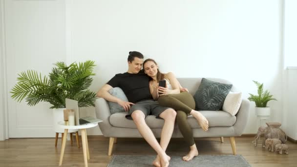 Ein Mann und eine Frau sitzen allein auf der Couch, halten eine Flasche Wasser in den Händen und schauen nach einem anstrengenden Arbeitstag gemeinsam auf das Telefon. — Stockvideo