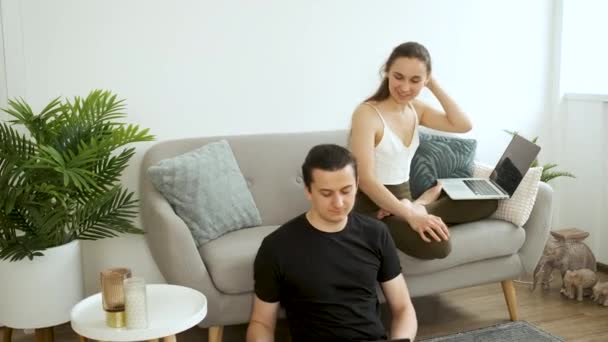 年轻夫妇坐在沙发上和地板上沉思，在笔记本电脑上工作。在家工作和冥想 — 图库视频影像