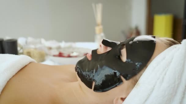 Una donna giace in una spa con una maschera nera sul viso. Ringiovanimento cutaneo e salute — Video Stock