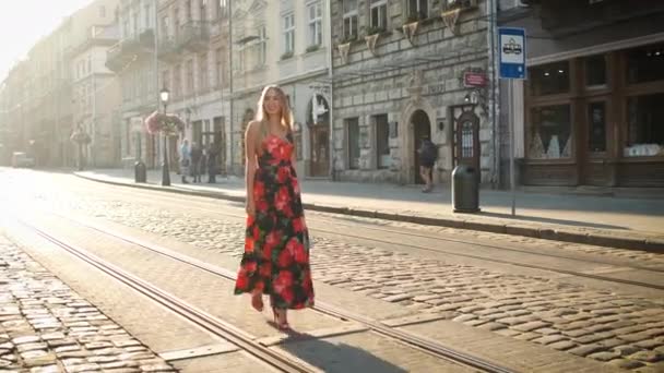 Vackra blonda kaukasiska utseende med ett uppriktigt leende och en vacker lång klänning med en strut promenader runt morgonstaden. Ställer sig framför kameran Tourist. Förlaga — Stockvideo