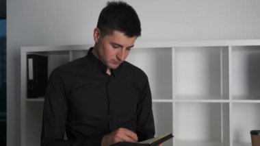 Genç bir adam 30 yaşında bir işadamı panoramik pencerelerle ofisinde duruyor ve bir defter işi fikirleri yazıyor.
