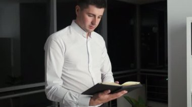Beyaz gömlekli beyaz tenli esmer, panoramik pencerenin yanında duruyor. Gece şehrine bakıyor ve bir kitap okuyor.