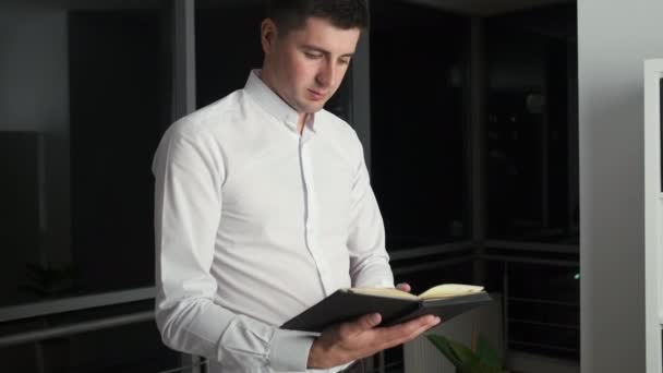 Beyaz gömlekli beyaz tenli esmer, panoramik pencerenin yanında duruyor. Gece şehrine bakıyor ve bir kitap okuyor. — Stok video