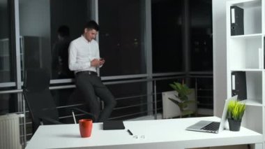 Ofiste panoramik pencerenin yanında elinde telefon tutan 30 yaşında bir adam var. Çevrimiçi arkadaşlarla sohbet