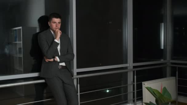 Pensive mladý muž 30 rok starý podnikatel v obleku stojící ve své kanceláři v blízkosti panoramatické okno s výhledem na noční město — Stock video