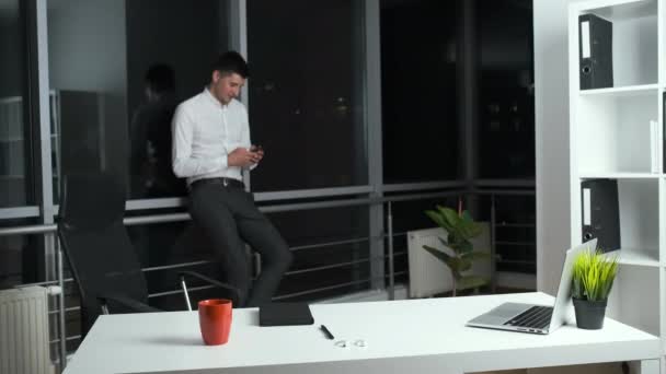 Třicetiletý muž stojí u panoramatického okna v kanceláři a v ruce drží telefon. Chat s přáteli online — Stock video
