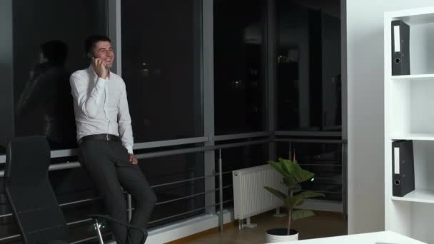 Ein 30-jähriger Mann steht im Büro vor einem Panoramafenster, hält ein Telefon in der Hand und kommuniziert mit Kunden. Arbeit bis spät in die Nacht — Stockvideo