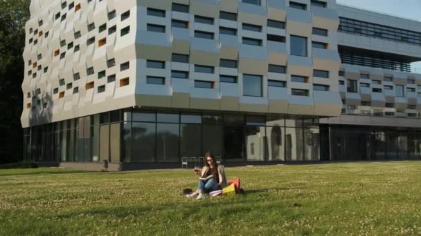 Gözlüklü ve kahverengi pelerinli şık bir kız parkta çimlerin üzerinde oturur ve bir kitap okur ve bir bardak kırmızı şarap içer. Hediye paketleri. Güneşli yaz akşamı — Stok video