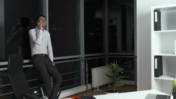 Třicetiletý muž stojí u panoramatického okna v kanceláři, v ruce drží telefon a komunikuje se zákazníky. Práce do pozdní noci — Stock video