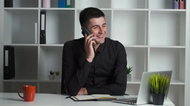 Un hombre de negocios de 30 años con el pelo negro en una camisa blanca se sienta en un escritorio en la oficina y se comunica con los clientes por teléfono. Administrador exitoso — Vídeos de Stock