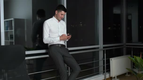Homem de 30 anos parado perto de uma janela panorâmica no escritório segurando um telefone em suas mãos. Converse com amigos online — Vídeo de Stock