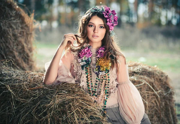 Attractive young woman with beautiful flowers near a haystack. Flowers in the hairstyle, costume jewelry, summer beauty concept