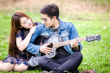 Asya çift çimen parkta oturan akustik şarkı gitar çalmak aşık