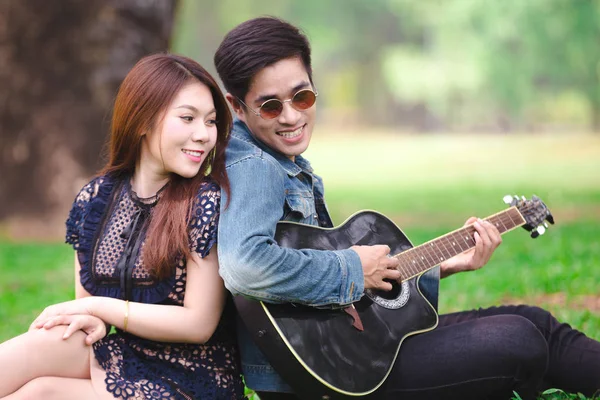 Asya çift çimen parkta oturan akustik şarkı gitar çalmak aşık