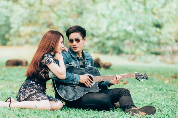 Asya çift çimen parkta oturan akustik şarkı gitar çalmak aşık