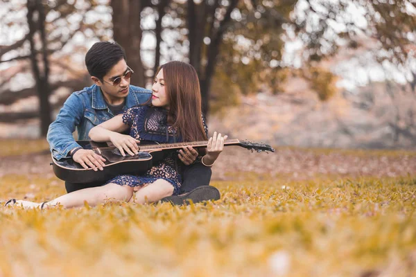 Asya çift çimen parkta oturan akustik şarkı gitar çalmak aşık