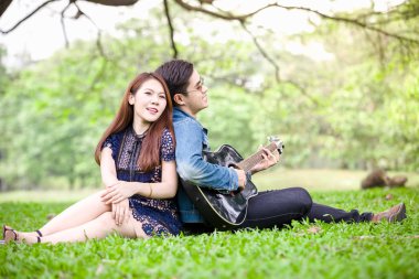 Asya çift çimen parkta oturan akustik şarkı gitar çalmak aşık