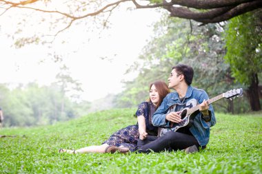 Asya çift çimen parkta oturan akustik şarkı gitar çalmak aşık
