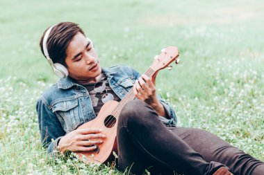 Asya genç erkek kulaklık ile müzik dinlemek doğa yalan ve ukulele oynamak