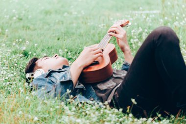 Asya genç erkek kulaklık ile müzik dinlemek doğa yalan ve ukulele oynamak
