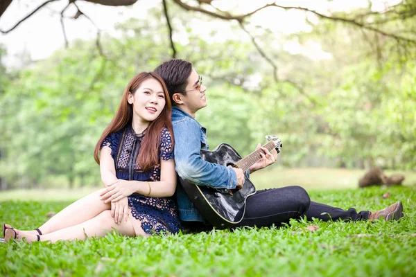 Asya çift çimen parkta oturan akustik şarkı gitar çalmak aşık