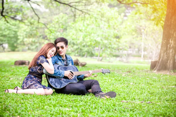 Asya çift çimen parkta oturan akustik şarkı gitar çalmak aşık