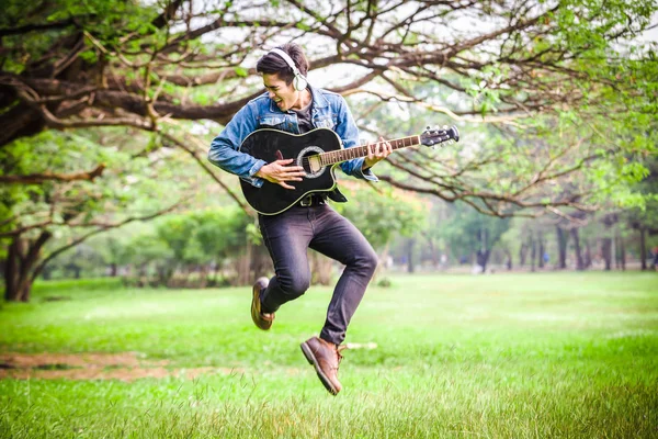 Asian young men jump in nature playing guitar and listen to music with headphones.