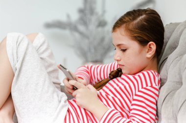 Kanepede oturan küçük kız oturma odasında akıllı telefon oynuyor.