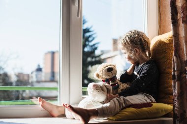 Pencerede otururken gün ışığının tadını çıkaran tatlı küçük kız. Çocuk büyük bir pencere pervazında oturuyor. D vitamini