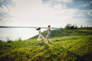 Sevgi dolu bir çift zıplıyor ve birbirlerinin elini tutuyor. Nehir ve gökyüzünün zeminine karşı. Çelenkli güzel bir kız ve şapkalı şık bir adam güneşli bir günde dışarıda vakit geçirir. Aşk hikayesi..