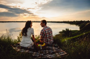 Nehir kıyısında gün batımında şarap içen sevgi dolu bir çift. Bir erkek arkadaş ve bir kız romantik bir randevuda, meyve sepetinin yanında oturup bardaktan şampanya içiyorlar..