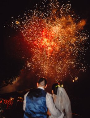 Gelin ve damat, gece gökyüzündeki güzel, renkli havai fişeklere bakarak kucaklaşıyorlar..