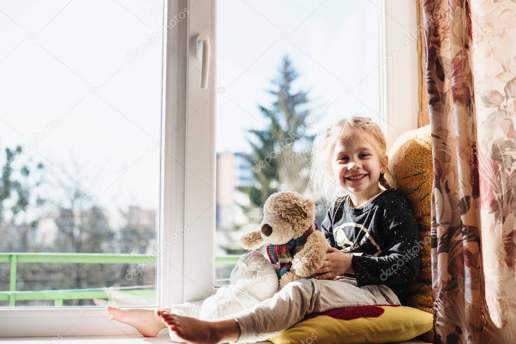 Cute little girl enjoying the sunshine while sitting at the window. The child is playing sitting on a large window sill. Vitamin D