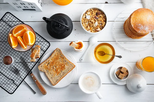 Концепция здорового завтрака, разнообразная утренняя еда - блинчики, яйцо мягкое, тосты, овсянка, мюсли, фрукты, кофе, чай, апельсиновый сок, молоко на белом деревянном столе — стоковое фото