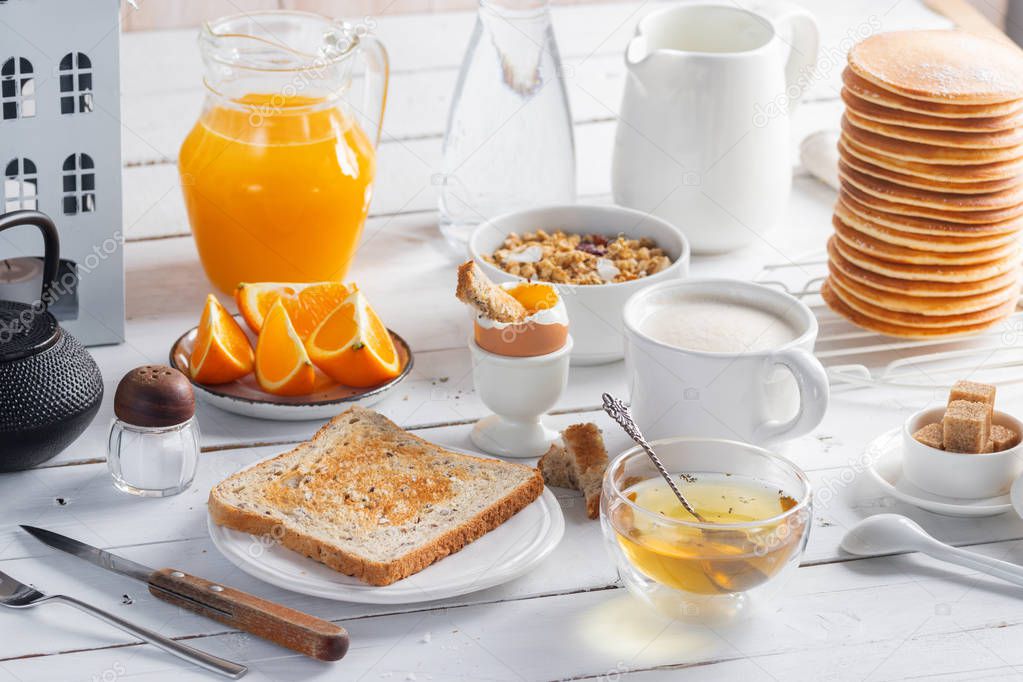 Healthy breakfast eating concept, various morning food - pancakes, soft-boiled egg, toast, oatmeal, granola, fruit, coffee, tea, orange juice, milk on white wooden table