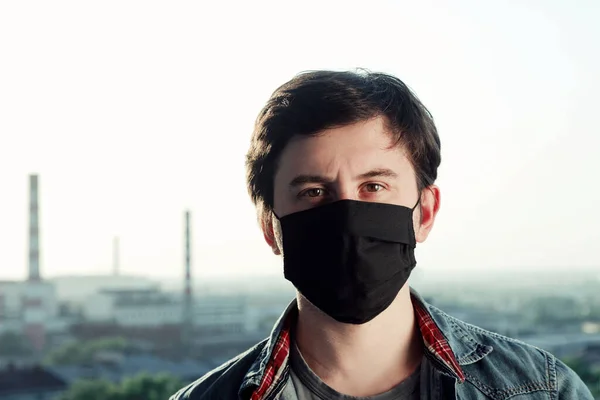 Молодий чоловік у чорній медичній масці та сорочці стоїть перед розмитим тлом індустріального міста — стокове фото