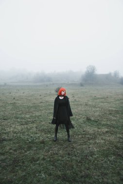 Kırmızı kısa saçlı, siyah elbiseli, soğuk, sisli sonbahar tarlasında moda kızı.
