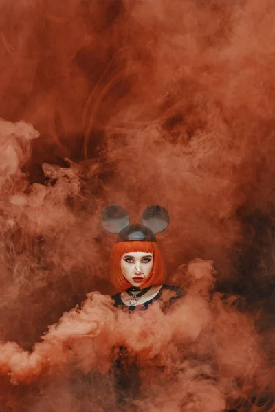Portrait of pretty fashion girl with red bob haircut and mouse hat in thick red smoke, young female with mouse hat and smoke bomb with dark background