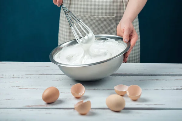 Woman in checkered apron whips egg whites on a white vintage wooden table. The process of making meringues.