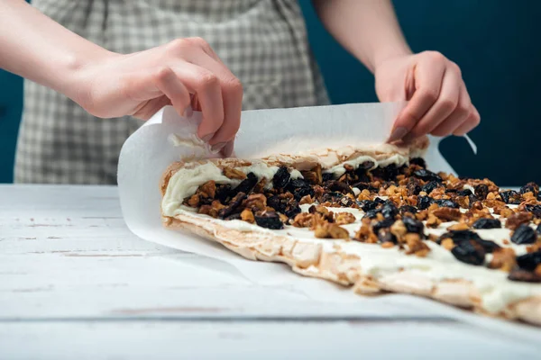 Woman in checkered apron rolls meringue roll on a white vintage wooden kitchen table. The process of making meringue roll.