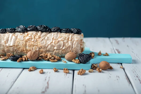 Prune-nut meringue roll on a blue cutting board on a white vintage wooden kitchen table. Meringue pie decorated with prunes and walnuts.