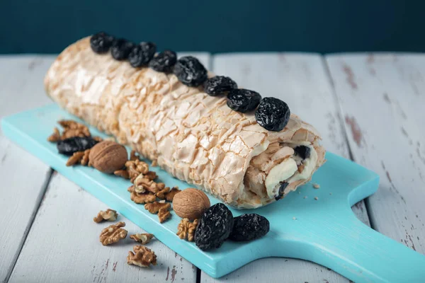 Prune-nut meringue roll on a blue cutting board on a white vintage wooden kitchen table. Meringue pie decorated with prunes and walnuts.