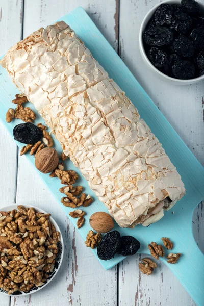 Prune-nut meringue roll on a blue cutting board on a white vintage wooden kitchen table. Meringue pie and bowls with prunes and walnuts.